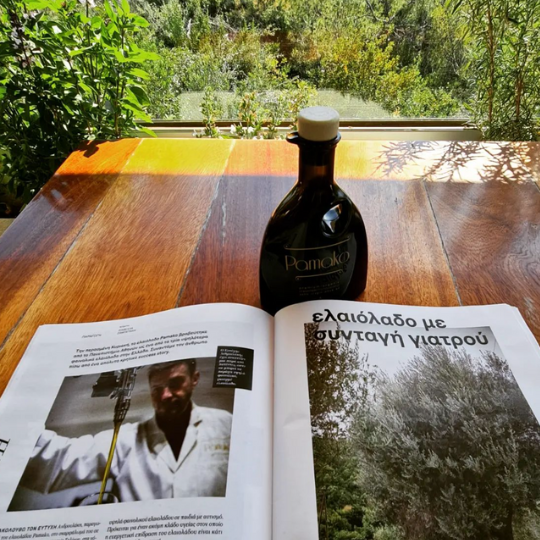 Bottle of Pamako olive oil beside an open magazine on a wooden table overlooking a green olive grove
