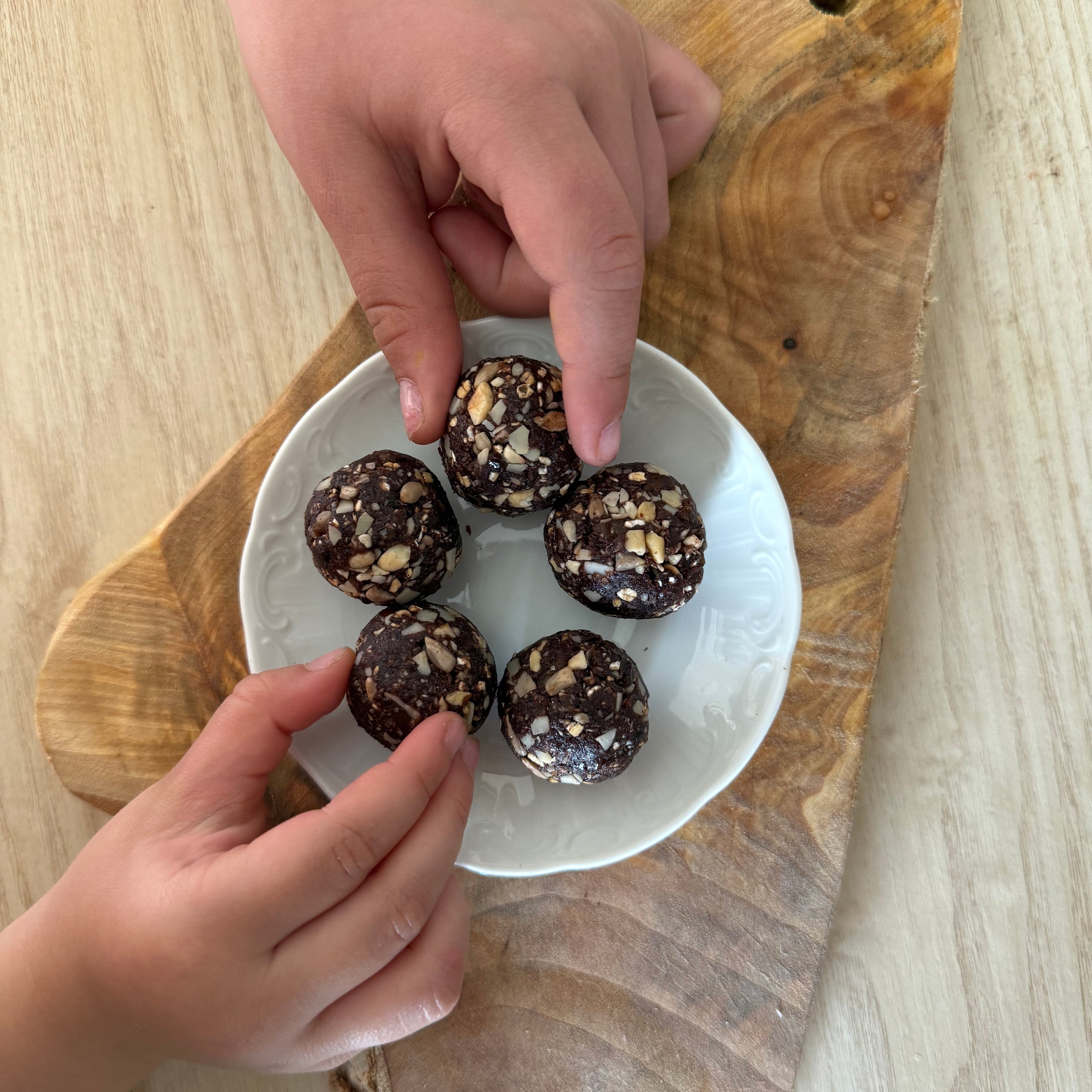 Five chocolate energy balls coated with chopped nuts on a white plate over a rustic wooden board, with hands reaching in