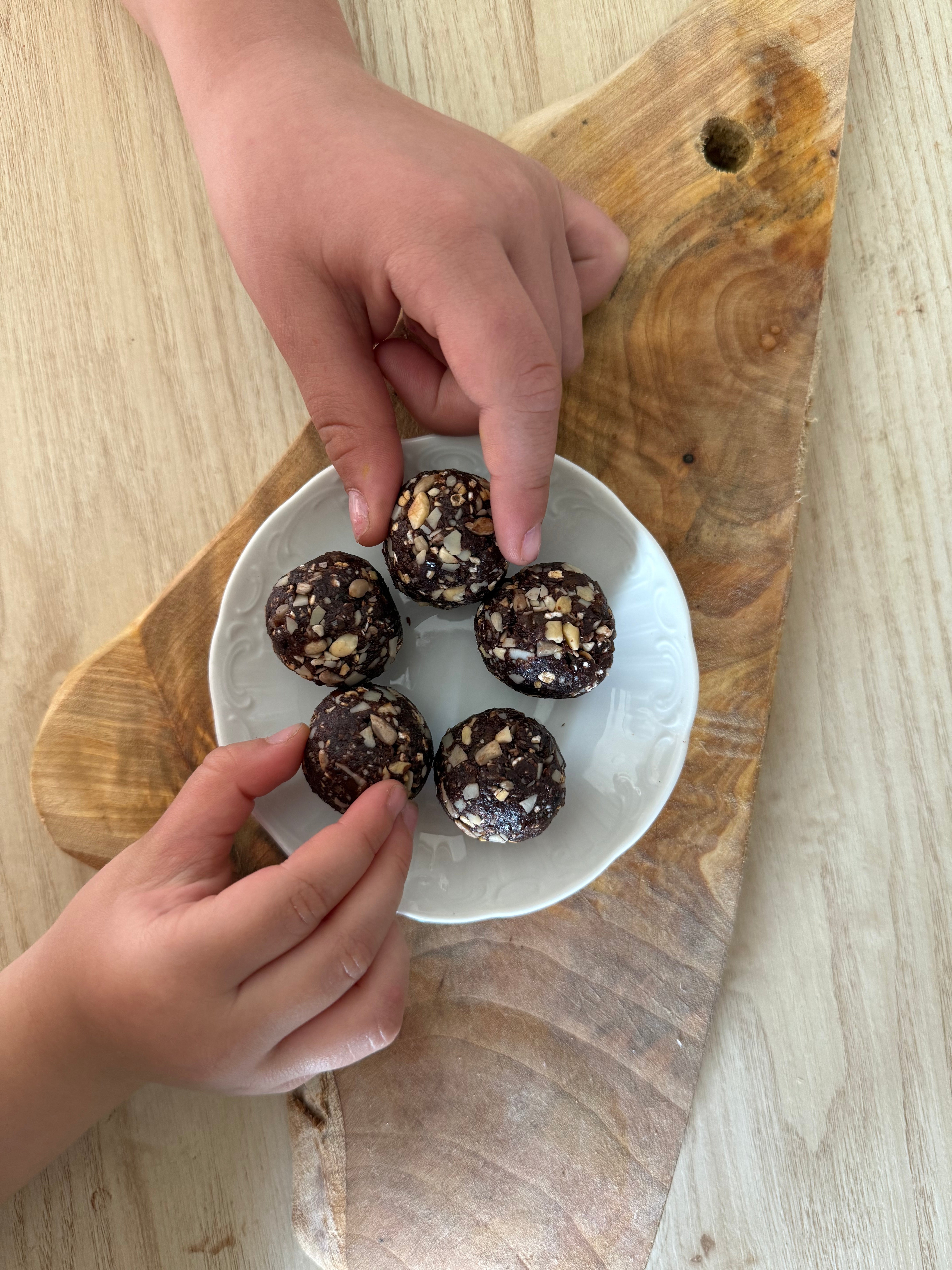 Five chocolate energy balls coated with chopped nuts on a white plate over a rustic wooden board, with hands reaching in